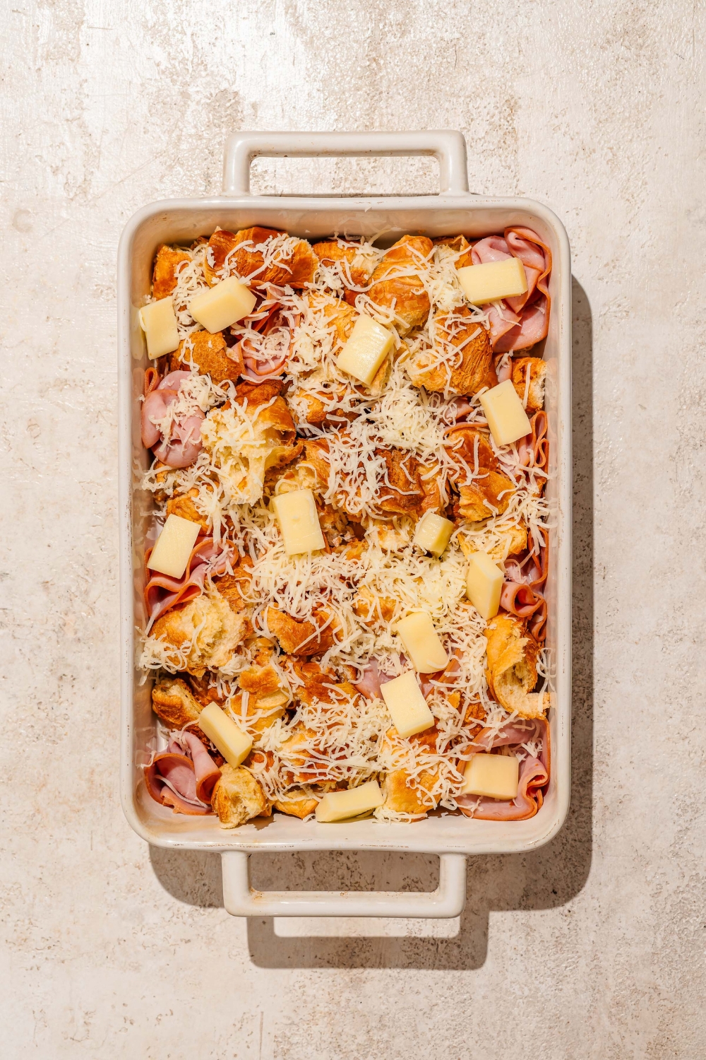 A baking dish with layers of sliced croissant, ham, and cheese. The dish is on a tan counter.
