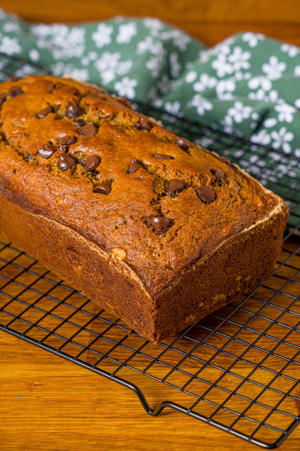 A loaf of baked chocolate chip banana bread with olive oil on a wire rack on a wooden counter with a green floral napkin.
