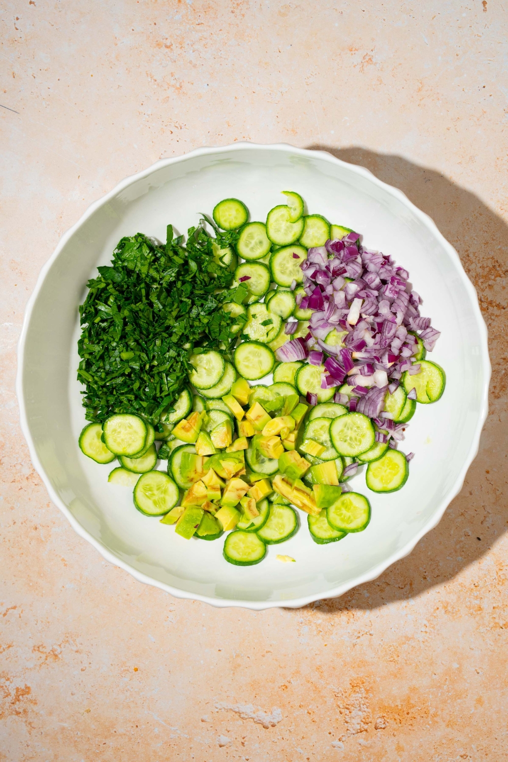 A large bowl with sliced cucumber, avocado, red onion, and fresh herbs. The bowl is on a tan counter.