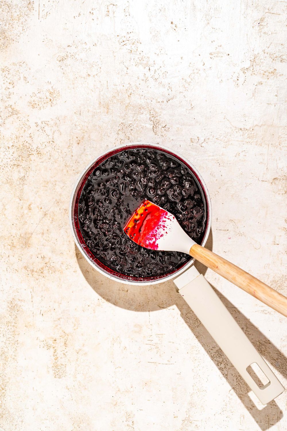 A saucepan with a spatula stirring a cooked elderberry jam mixture. The pot is on a tan counter.