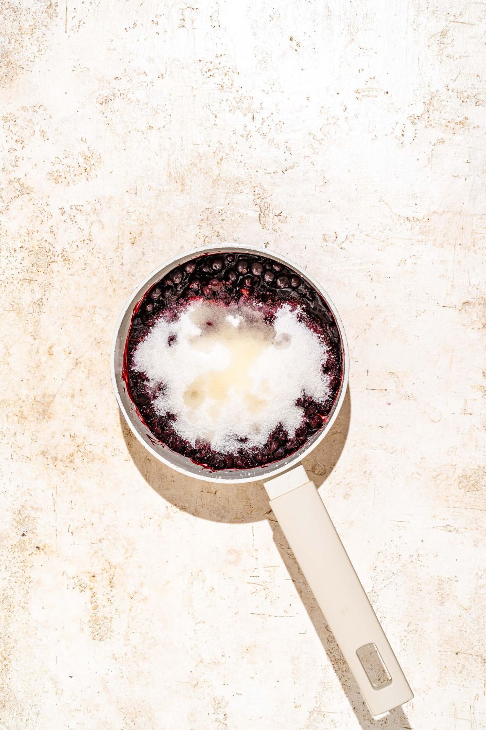A saucepan with crushed elderberries topped with sugar and lemon juice. The pot is on a tan counter.