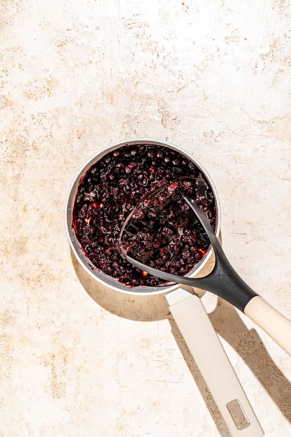 A saucepan with elderberries and a masher mashing the berries. The pot is on a tan counter.