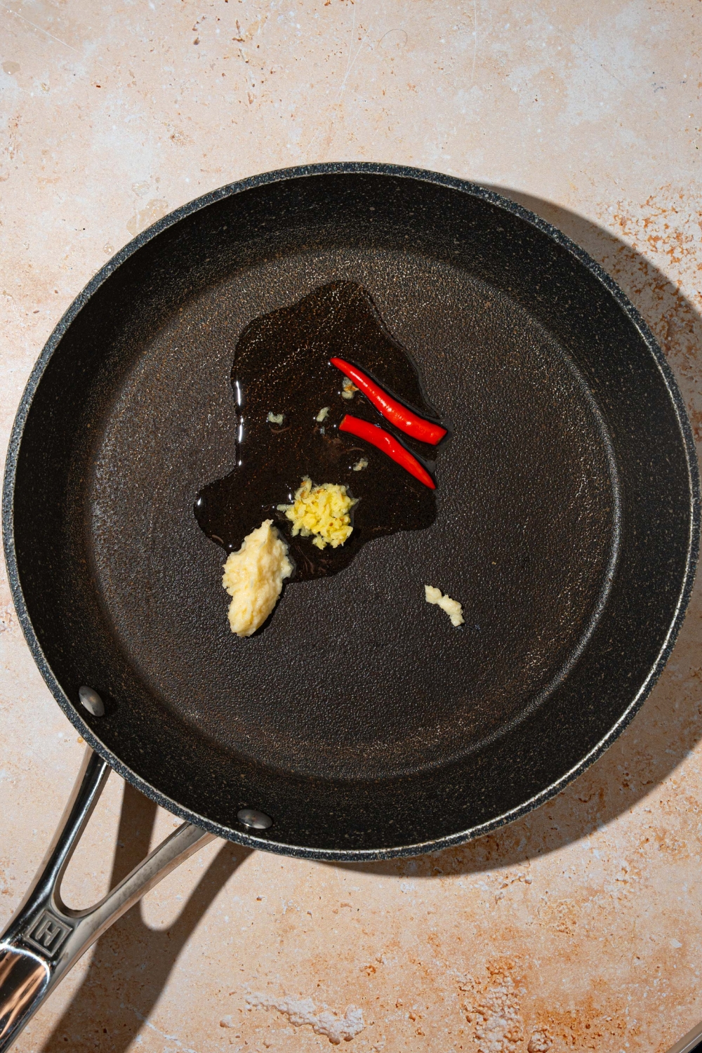 A skillet with red chiles, garlic, and ginger cooking in oil. The skillet is on a tan counter.
