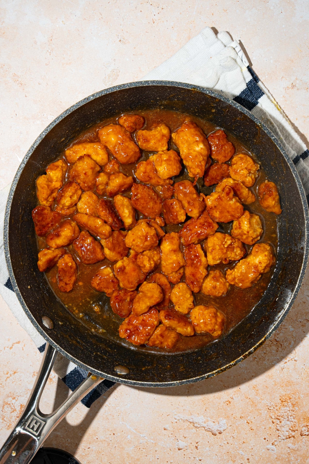 A skillet with crispy chicken tossed in general tso sauce. The skillet is on a tan counter.