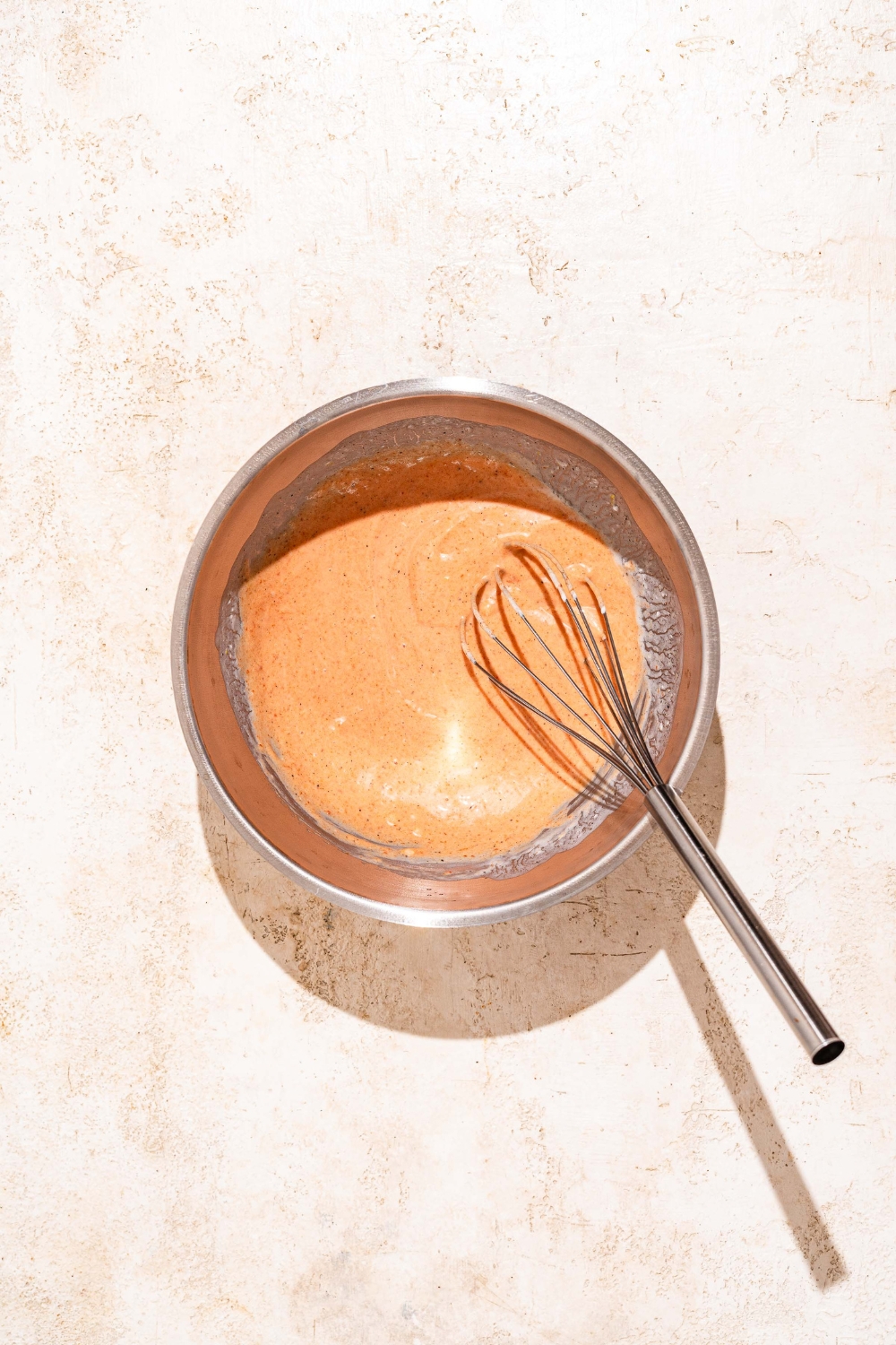 A large bowl with a whisk mixing ingredients to make gochujang mayo. The bowl is on a tan counter.
