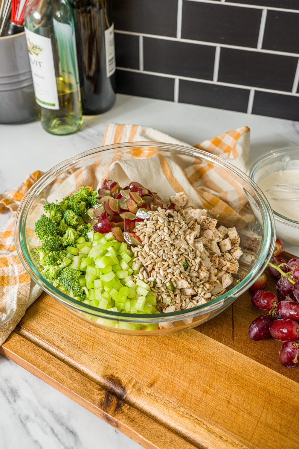 A large glass bowl with ingredients to make broccoli chicken salad including broccoli, chicken, grapes, celery, and sunflower seeds. The bowl is on a wooden board with grapes and a bowl of dressing.