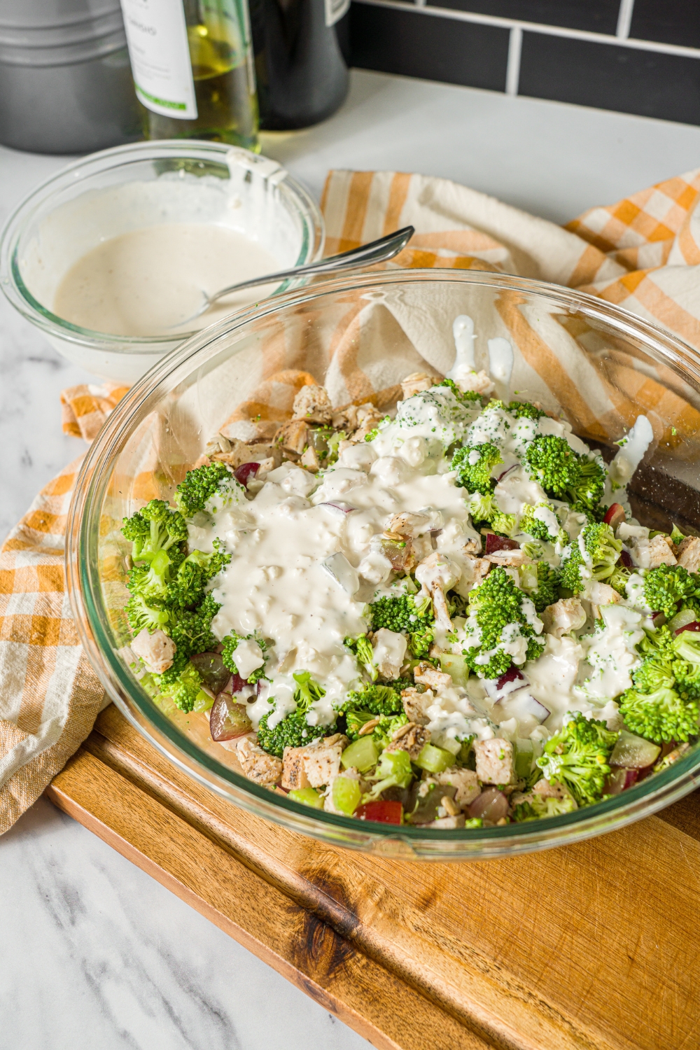 A large bowl with broccoli, chicken, grapes, and celery topped with a greek yogurt dressing. The bowl is on a wooden board with a yellow checkered napkin and small bowl of dressing.