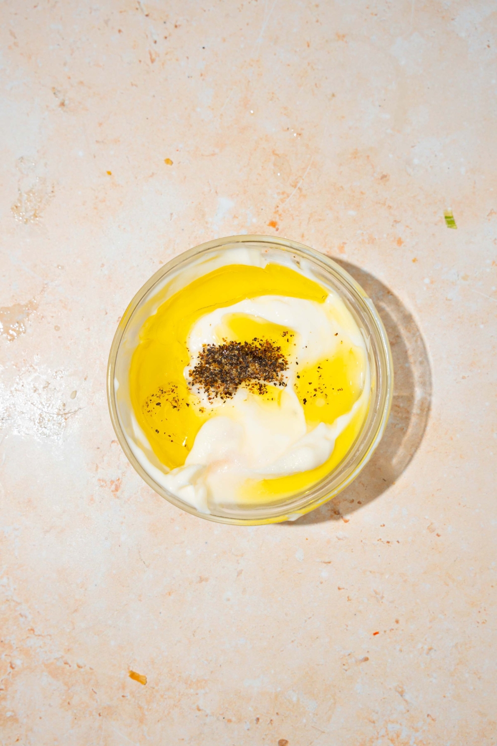 A small bowl with ingredients to make greek yogurt dressing including greek yogurt, oil, and seasonings. The bowl is on a tan counter.