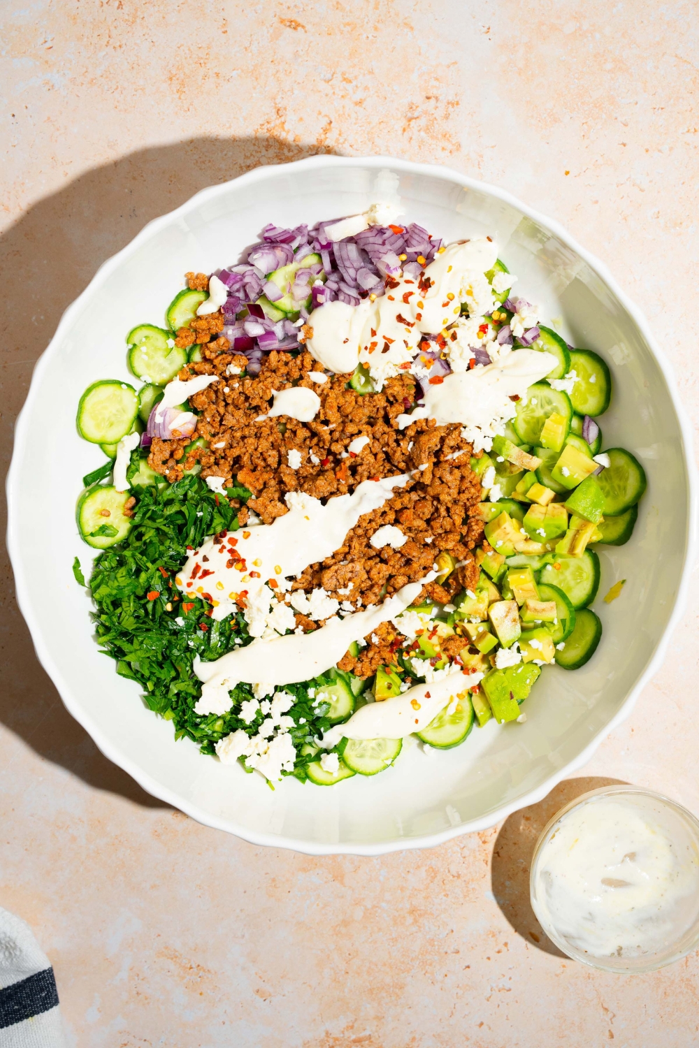 A large bowl with sliced cucumber, red onion, seasoned ground beef, avocado, and fresh herbs drizzled with greek yogurt dressing. The bowl is on a tan counter.