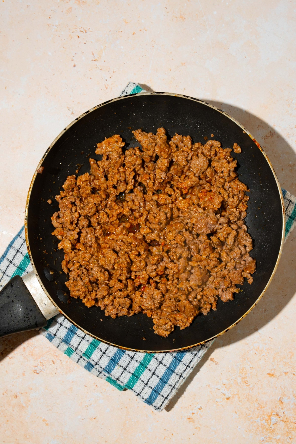 A skillet with cooked seasoned ground beef. The skillet is on a blue checkered napkin on a tan counter.