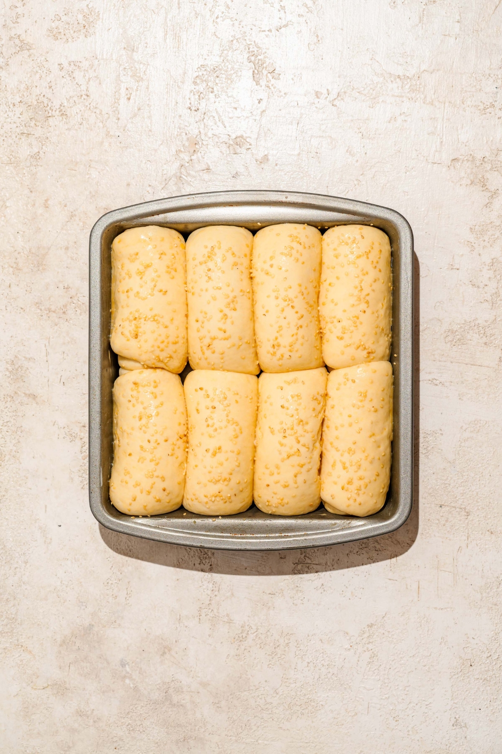 A large loaf pan with several small loaves of Raising Cane's bread dough brushed with egg wash and topped with sesame seeds. The pan is on a tan counter.