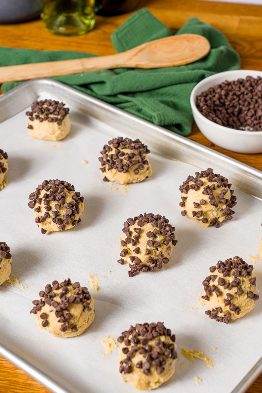 A baking sheet lined with parchment paper with chocolate chip cookie dough balls made without baking soda rolled in mini chocolate chips. The sheet is on a wooden counter with a bowl of chocolate chips.