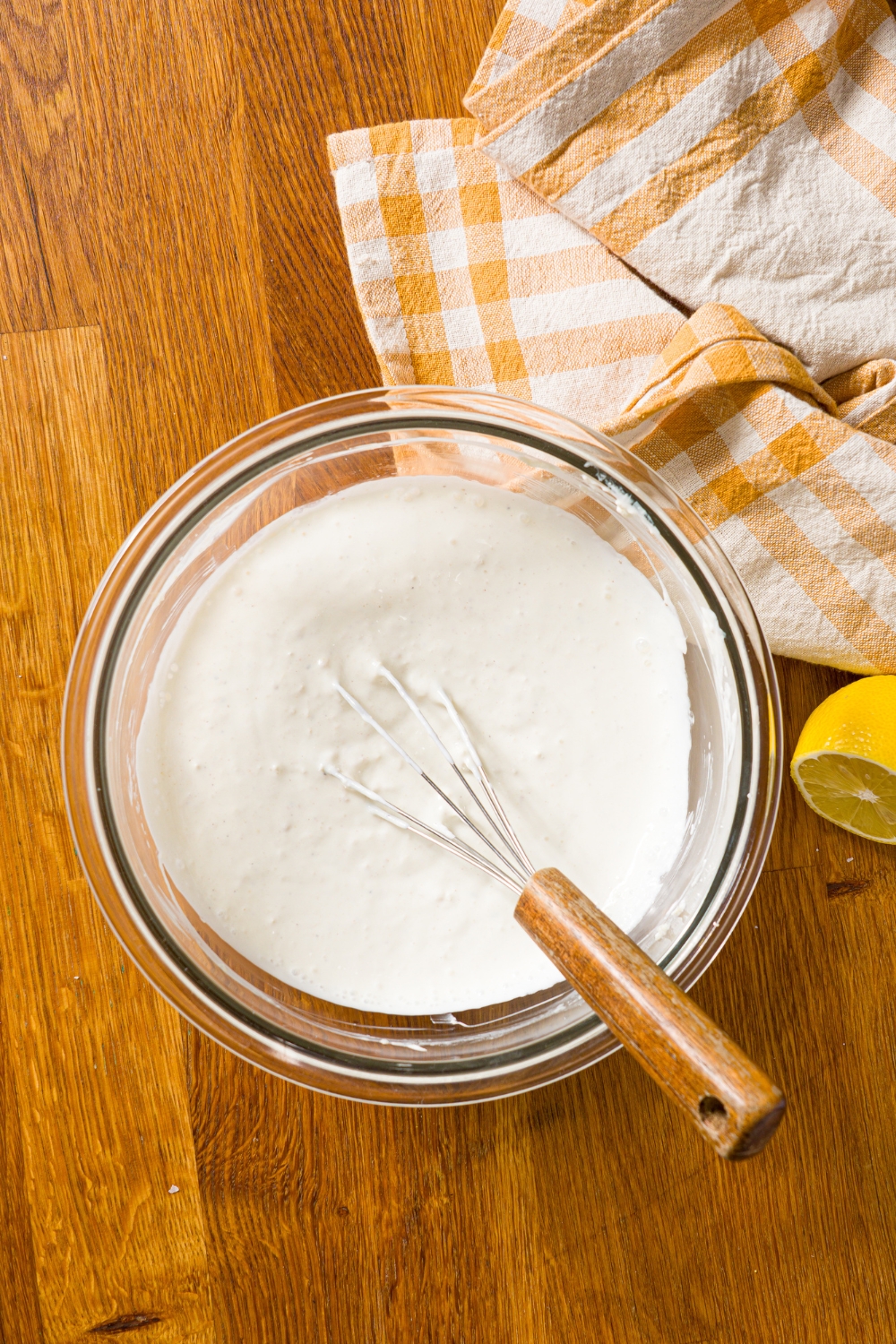 A glass bowl with a whisk mixing ingredients to make creamy horseradish sauce. The bowl is on a wooden counter with a yellow checkered napkin.