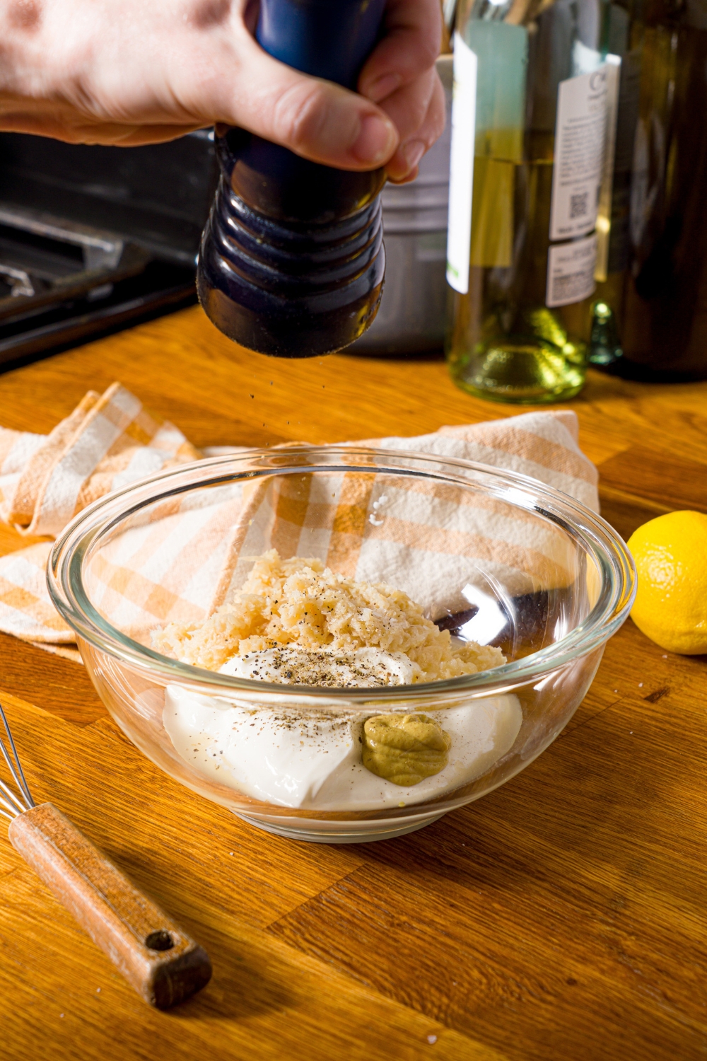 A glass bowl with ingredients to make horseradish sauce including sour cream, horseradish, mustard, with pepper being added to the bowl. The bowl is on a wooden counter with a yellow checkered napkin.