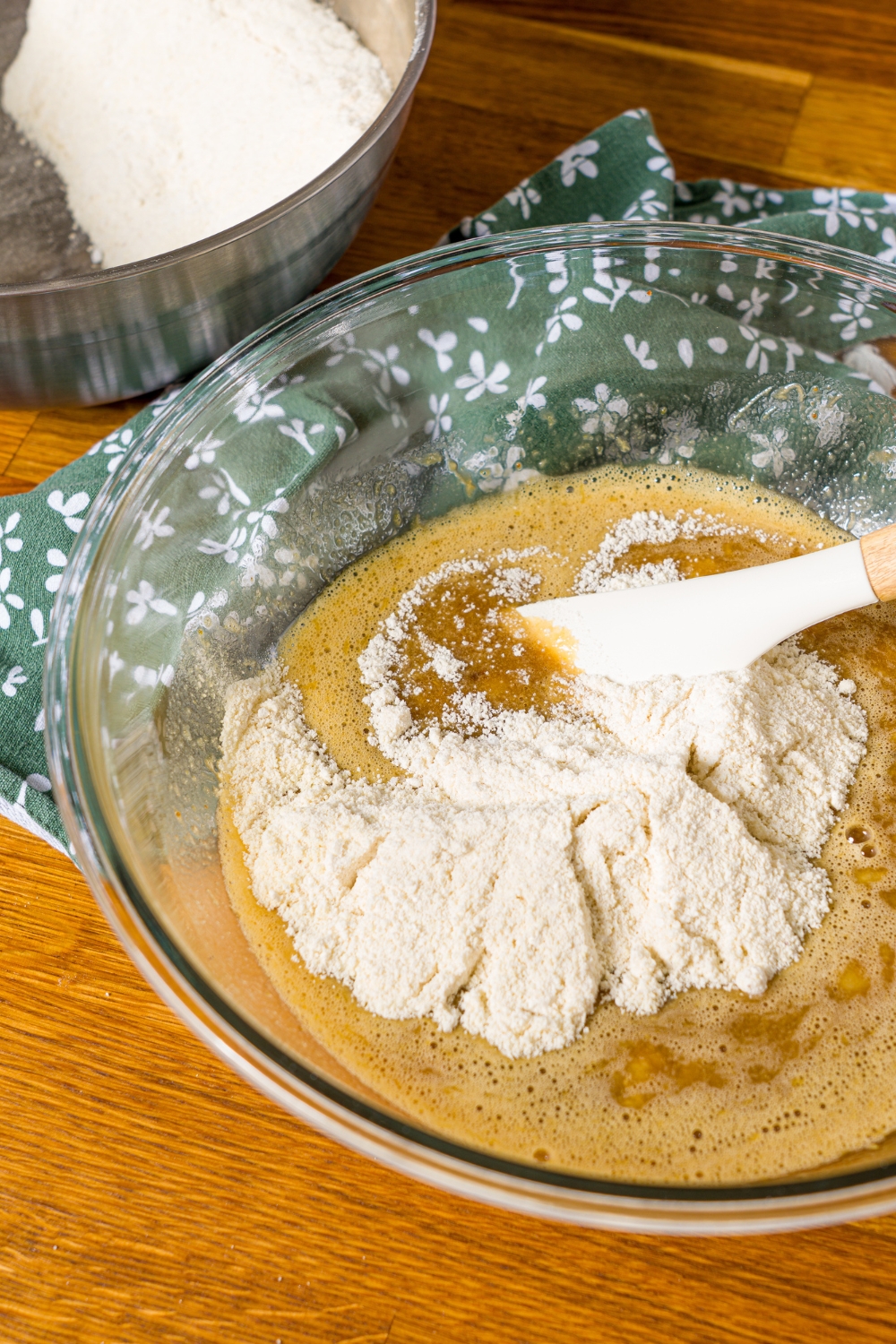 A large bowl with a wet banana bread batter mixture topped with dry ingredients. A spatula is combining the ingredients. The bowl is on a wooden counter with a green floral napkin.