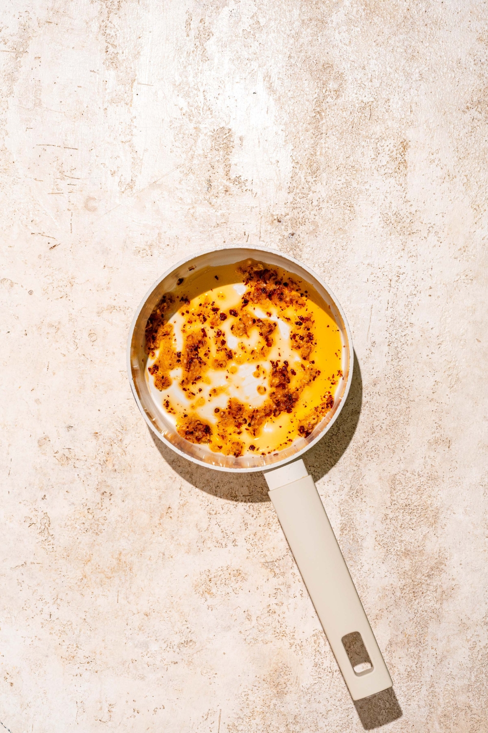 A saucepan with garlic, ginger, and chili flakes sautéing in oil. The saucepan is on a tan counter.