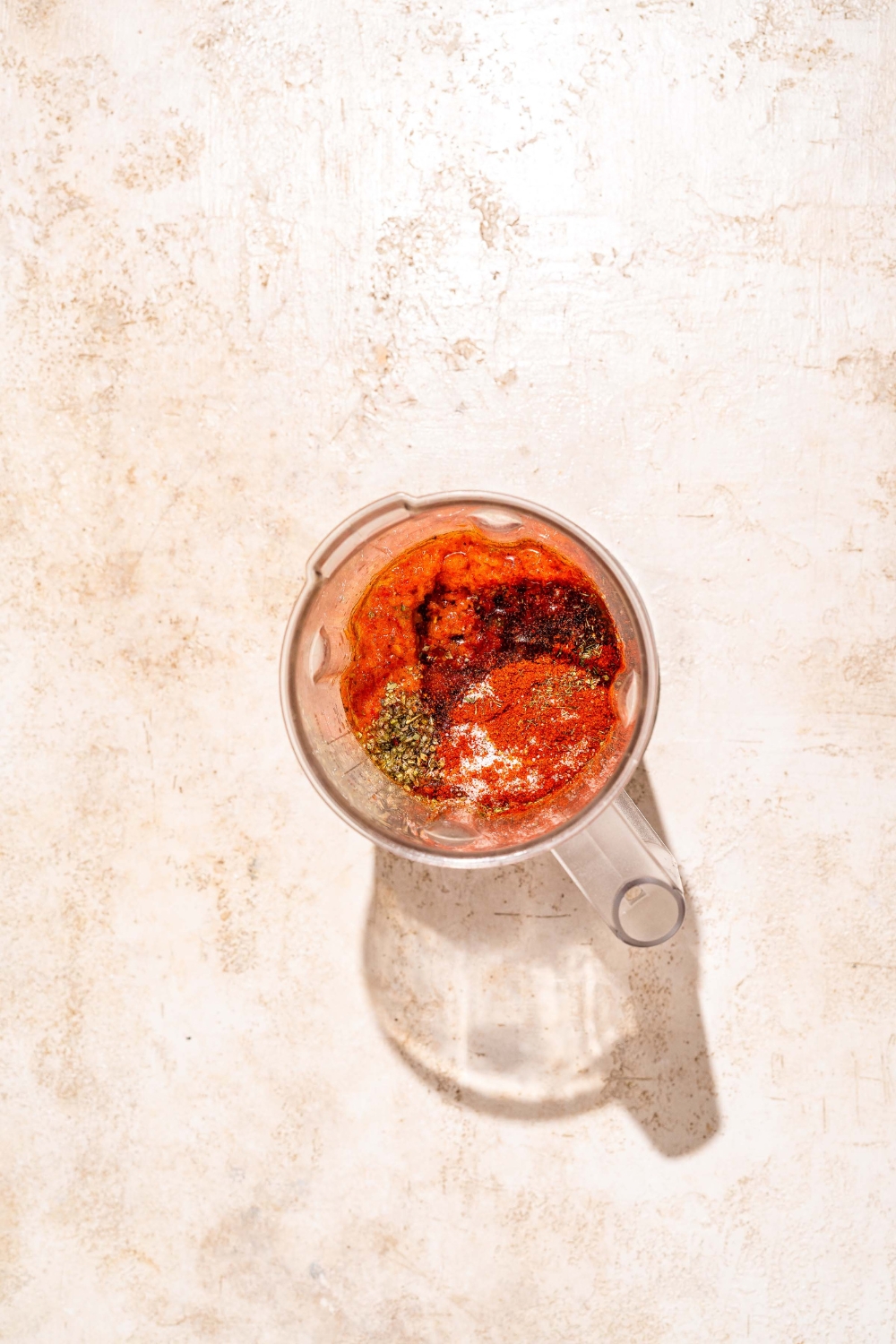 A blender jar with blended fresh ingredients topped with seasonings for peri peri sauce. The jar is on a tan counter.