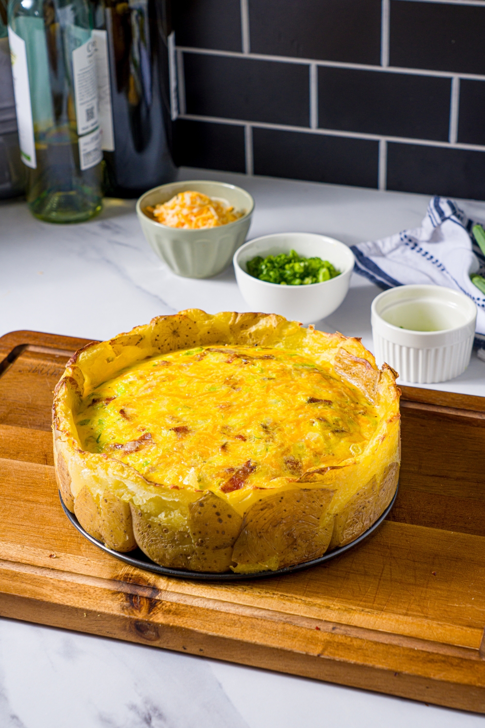 A wooden board with a potato crust quiche on an open springform pan. The board is on a marble counter with bowls of ingredients.