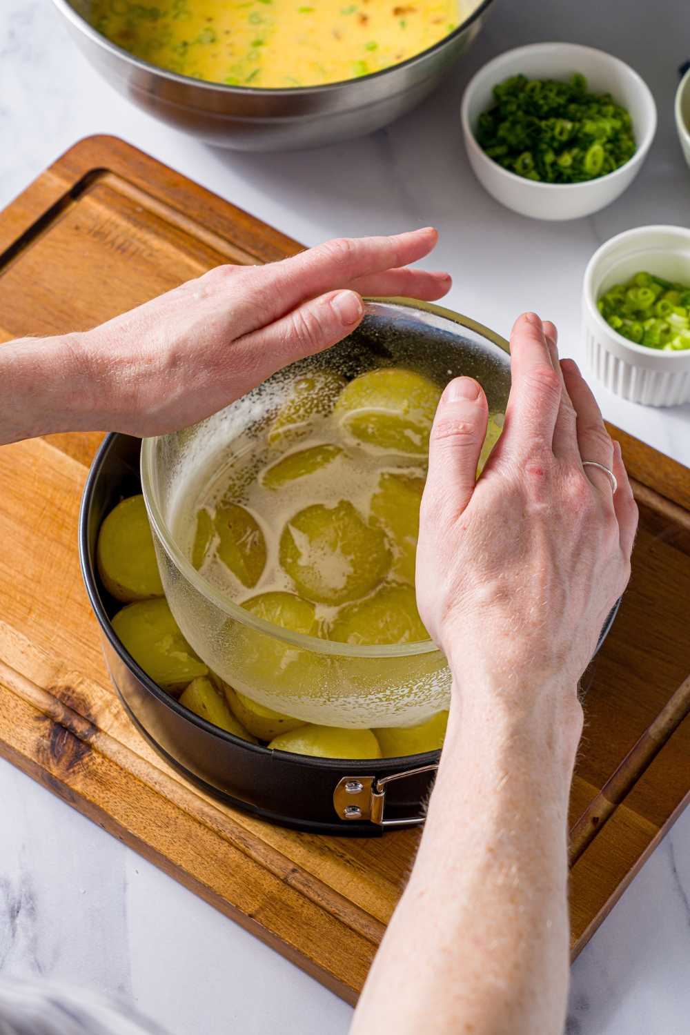 A glass bowl pressing cooked potatoes in a springform pan. The pan is on a wooden board on a marble counter with bowls of ingredients.