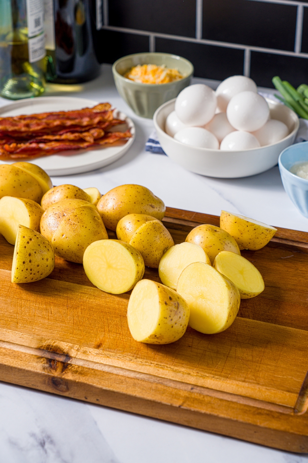 A wooden board with sliced potatoes. The board is on a marble counter with bowls of ingredients to make quiche.