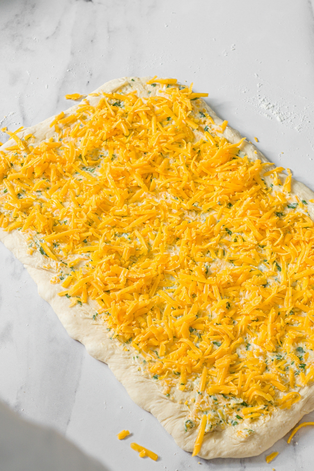 A marble counter with rolled out bread dough topped with a garlic mixture and topped with shredded cheese.