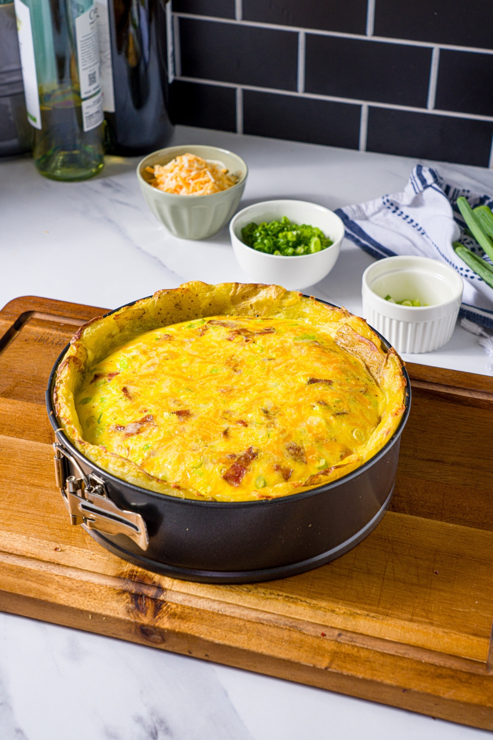 A wooden board with a baked potato crust quiche in a springform pan. The board is on a marble counter with small bowls of ingredients.
