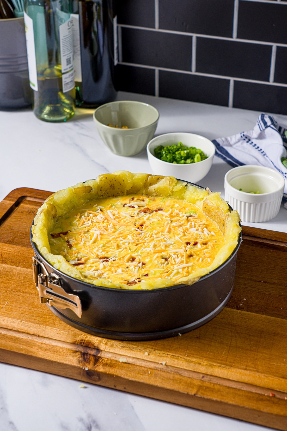 A wooden board with an unbaked potato crust quiche in a springform pan. The board is on a marble counter with small bowls of ingredients.