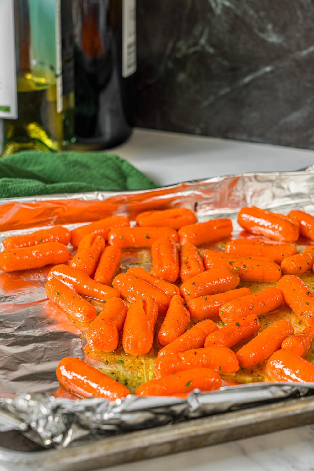 A baking sheet lined with foil with baby carrots coated in honey glaze and fresh parsley. The sheet is on a marble counter.