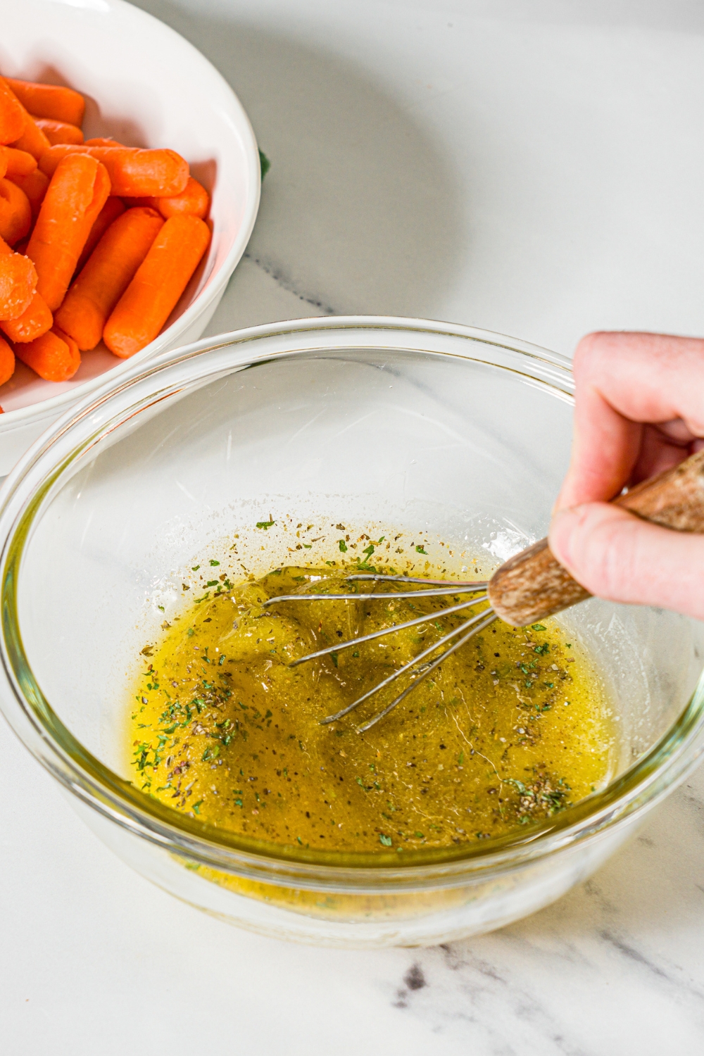 A small glass bowl with a whisk mixing a honey glaze including honey, oil, and seasonings. The bowl is on a marble counter with a bowl of baby carrots.