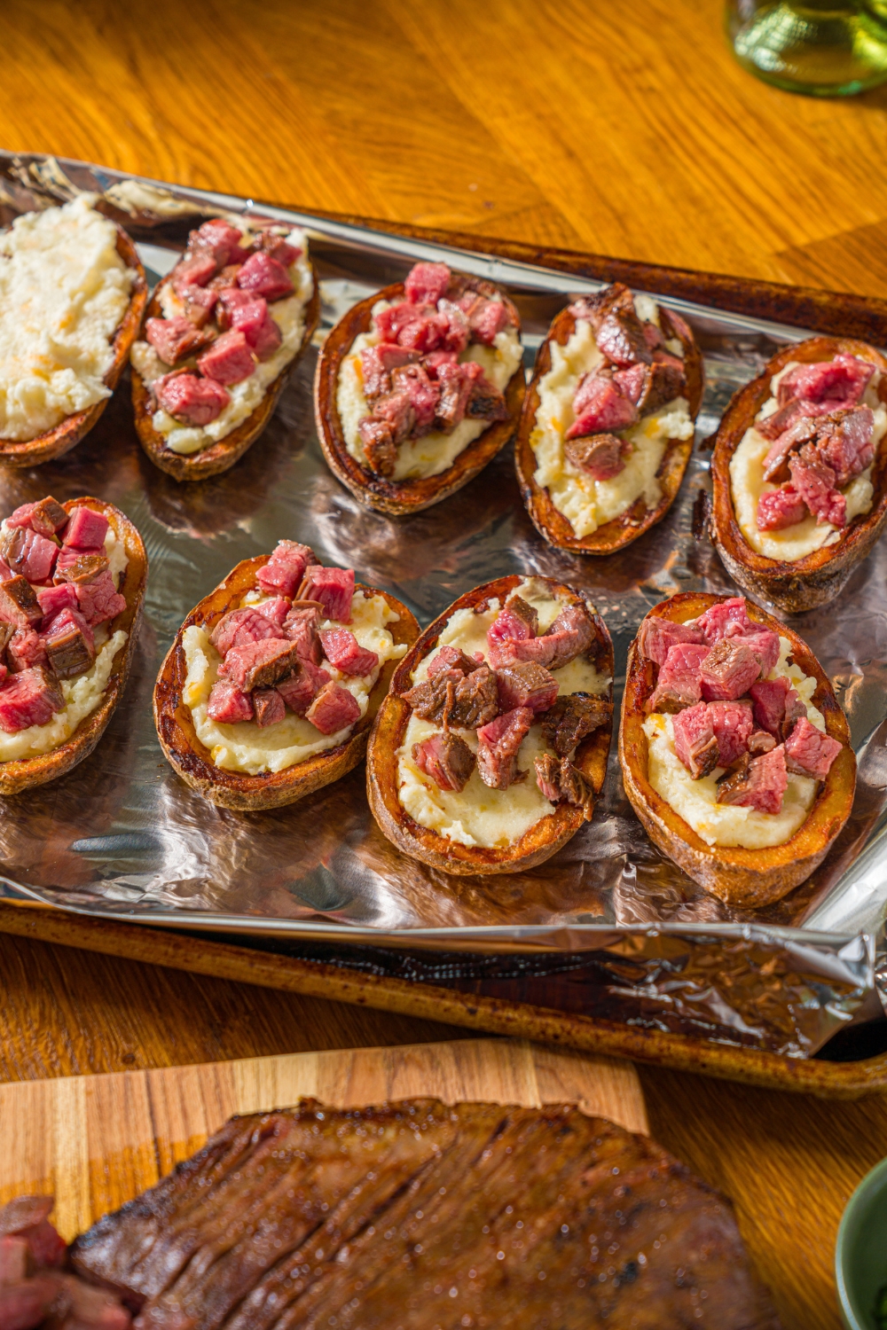 A baking sheet lined with foil with fried potato skins topped with potato filling and cubed steak. The sheet is on a wooden counter.