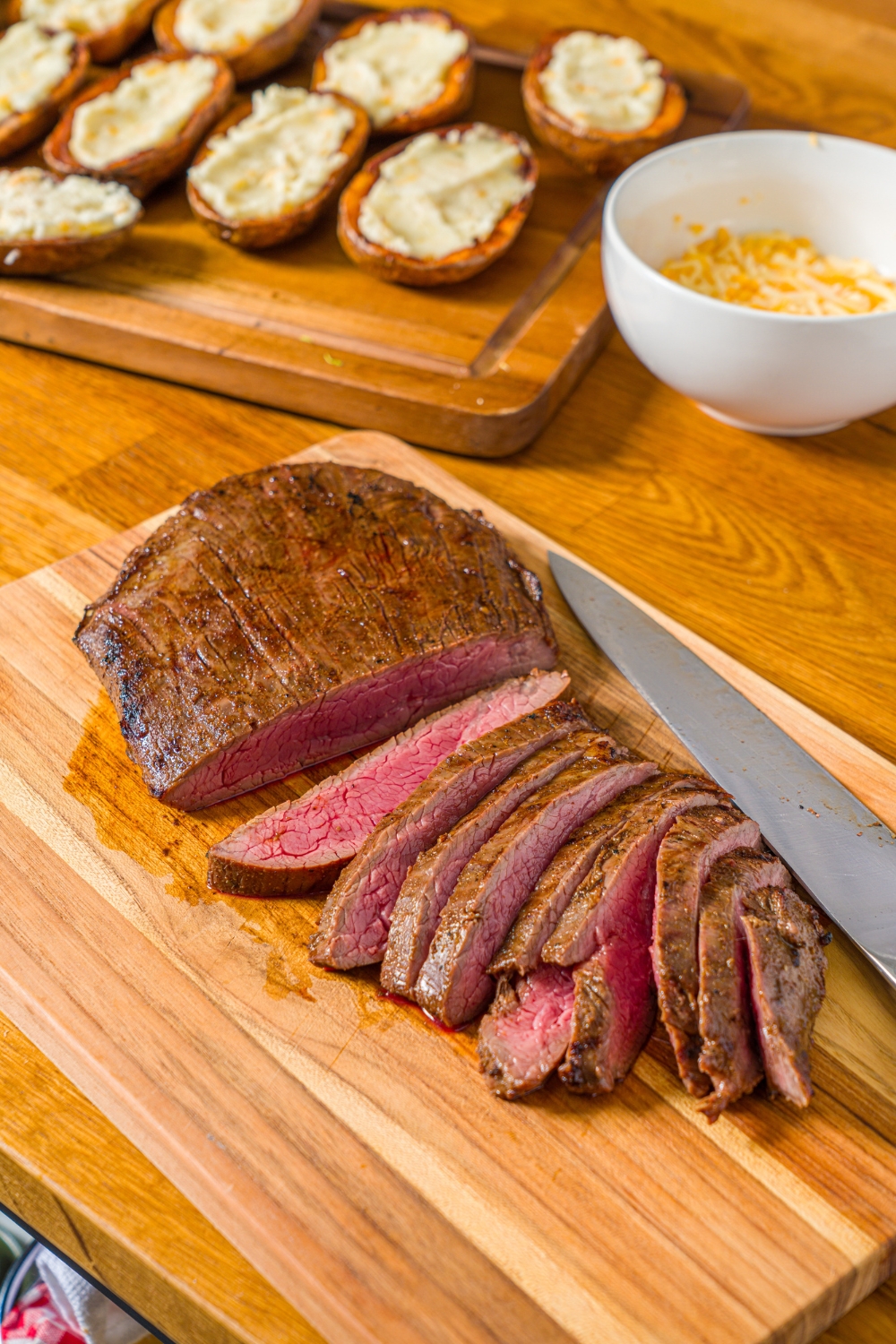 A wooden cutting board with a knife and sliced steak. The board is on a wooden counter with a board of stuffed potato skins.