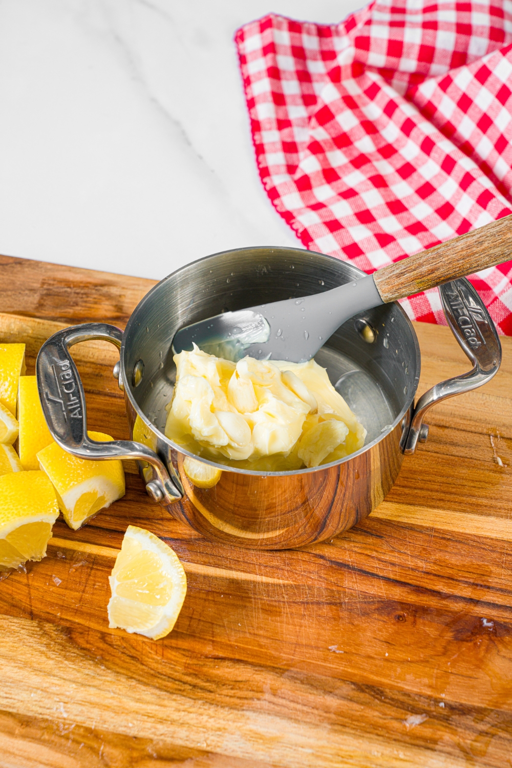 A small saucepan with a spatula mixing a butter and lemon sauce. The saucepan is on a wooden board with sliced lemon.