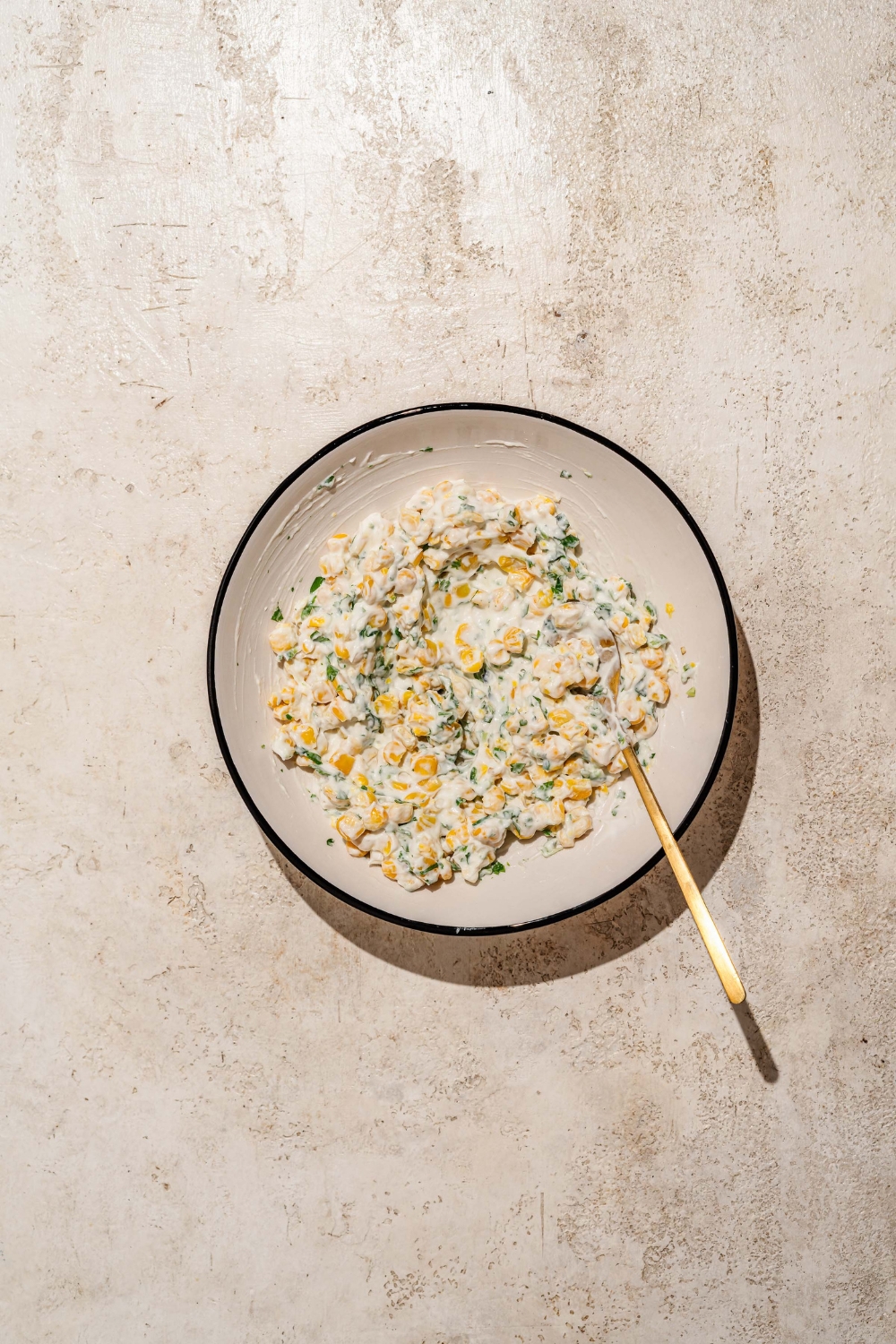 A bowl with a spoon mixing a street corn mixture including corn, jalapeño, cilantro, sour cream, mayo, and lime. The bowl is on a tan counter.