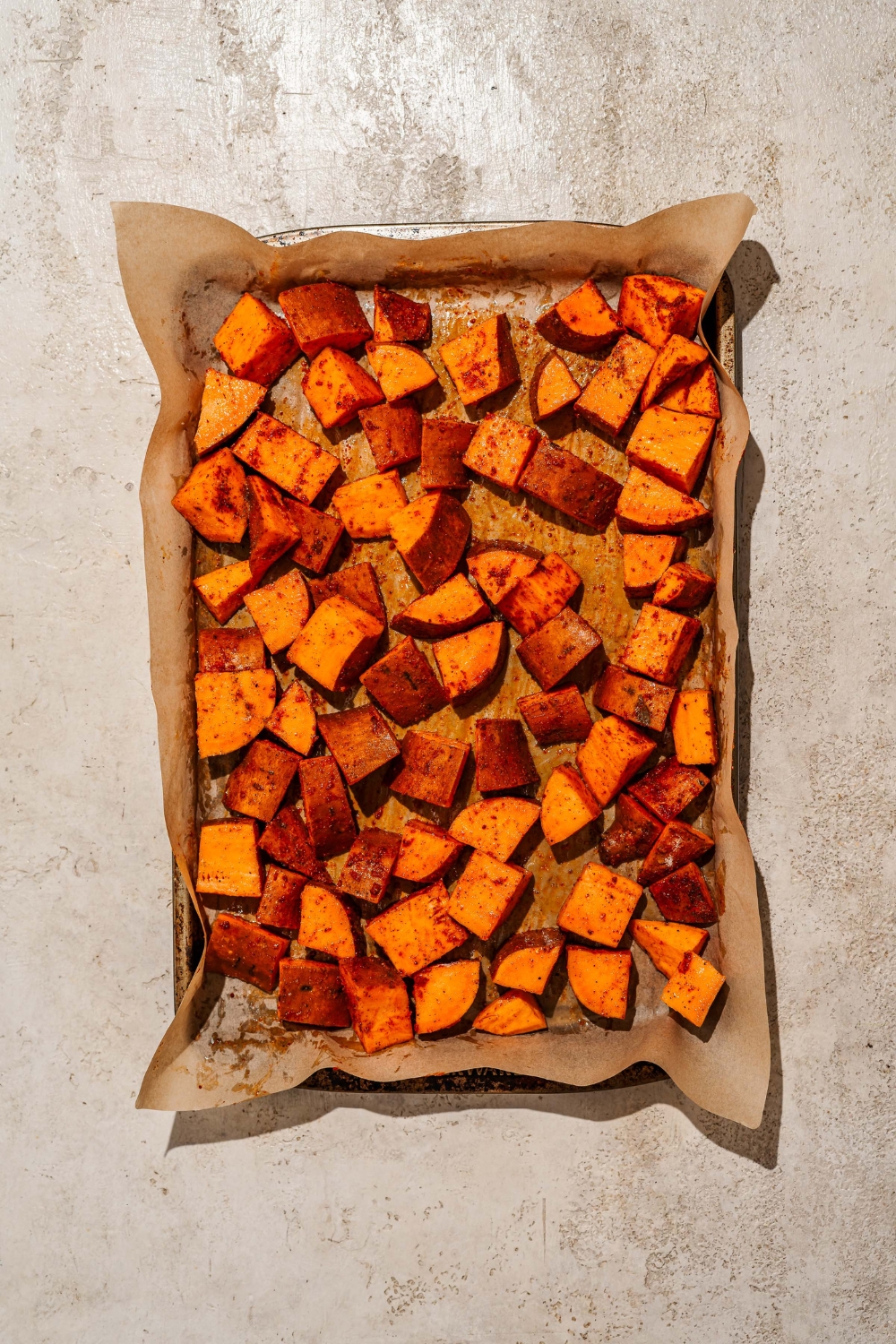 A baking sheet lined with parchment paper with seasoned roasted sweet potatoes. The baking sheet is on a tan counter.