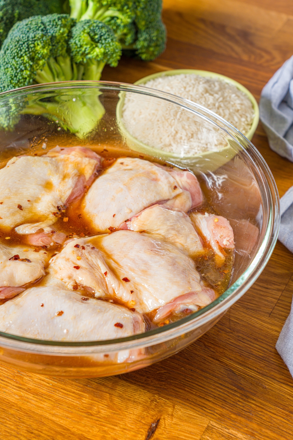 A large bowl with several chicken thighs marinating in a teriyaki marinade. The bowl is on a wooden counter with a bowl of rice and broccoli.