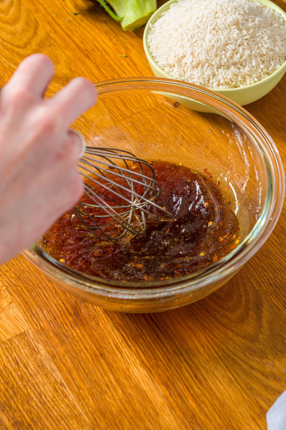 A bowl with a whisk mixing ingredients to make a teriyaki marinade. The bowl is on a wooden counter with a bowl of rice.