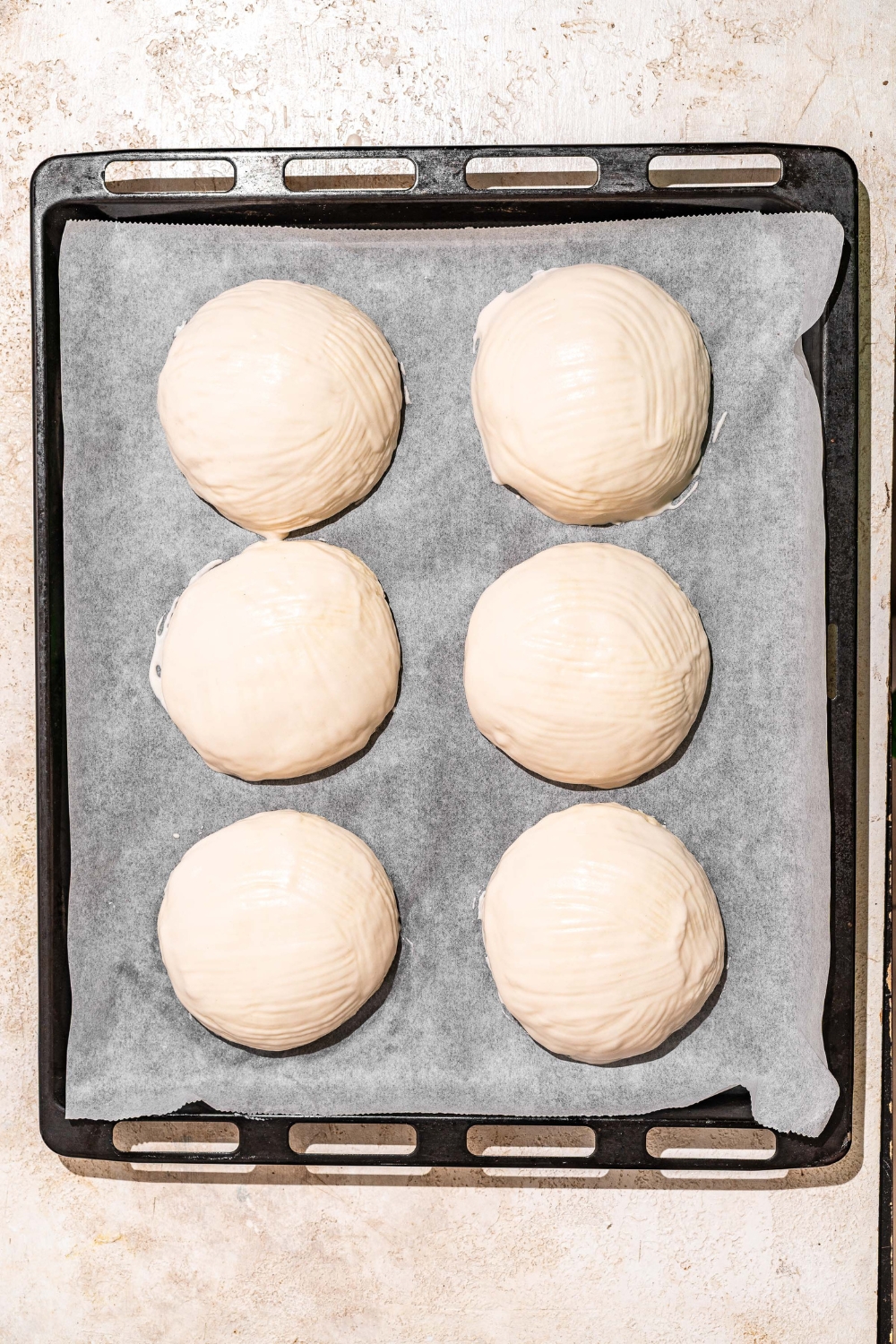A baking sheet lined with parchment paper with several unbaked loaves of Dutch Crunch bread dough. The sheet is on a tan counter.