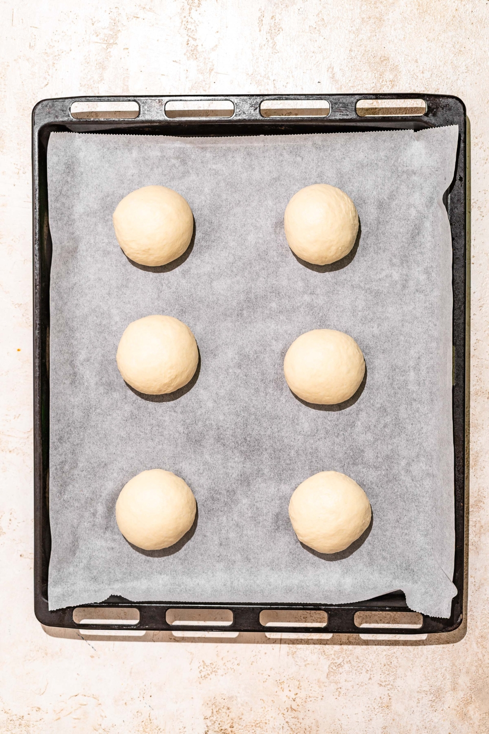A baking sheet lined with parchment paper with several unbaked loaves of Dutch Crunch dough balls. The sheet is on a tan counter.