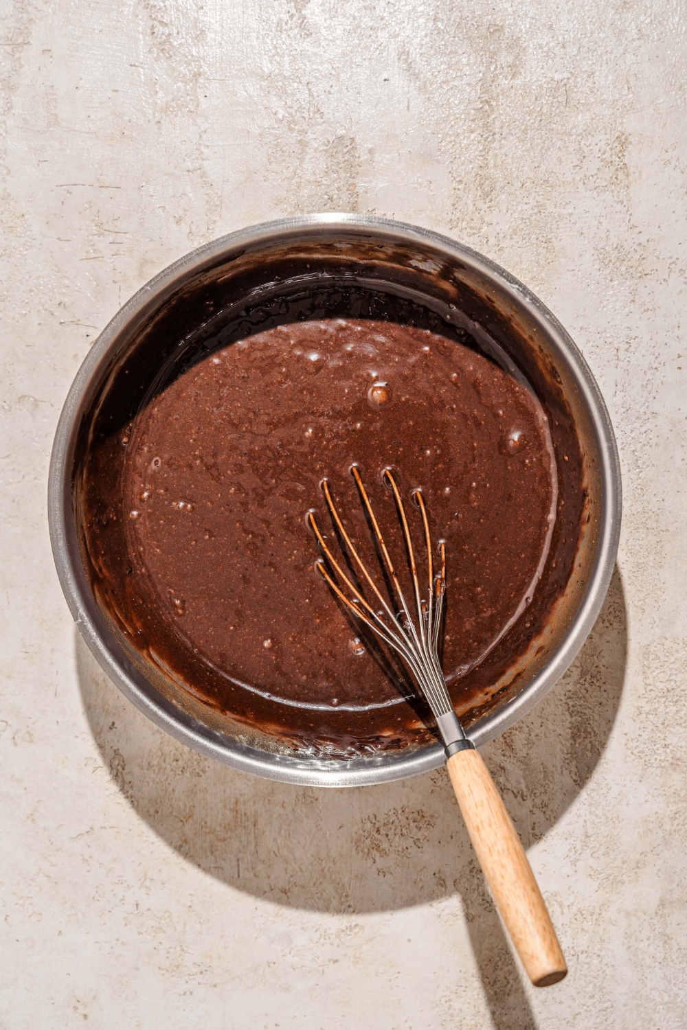 A large mixing board with a whisk mixing chocolate cake batter. The bowl is on a tan counter.