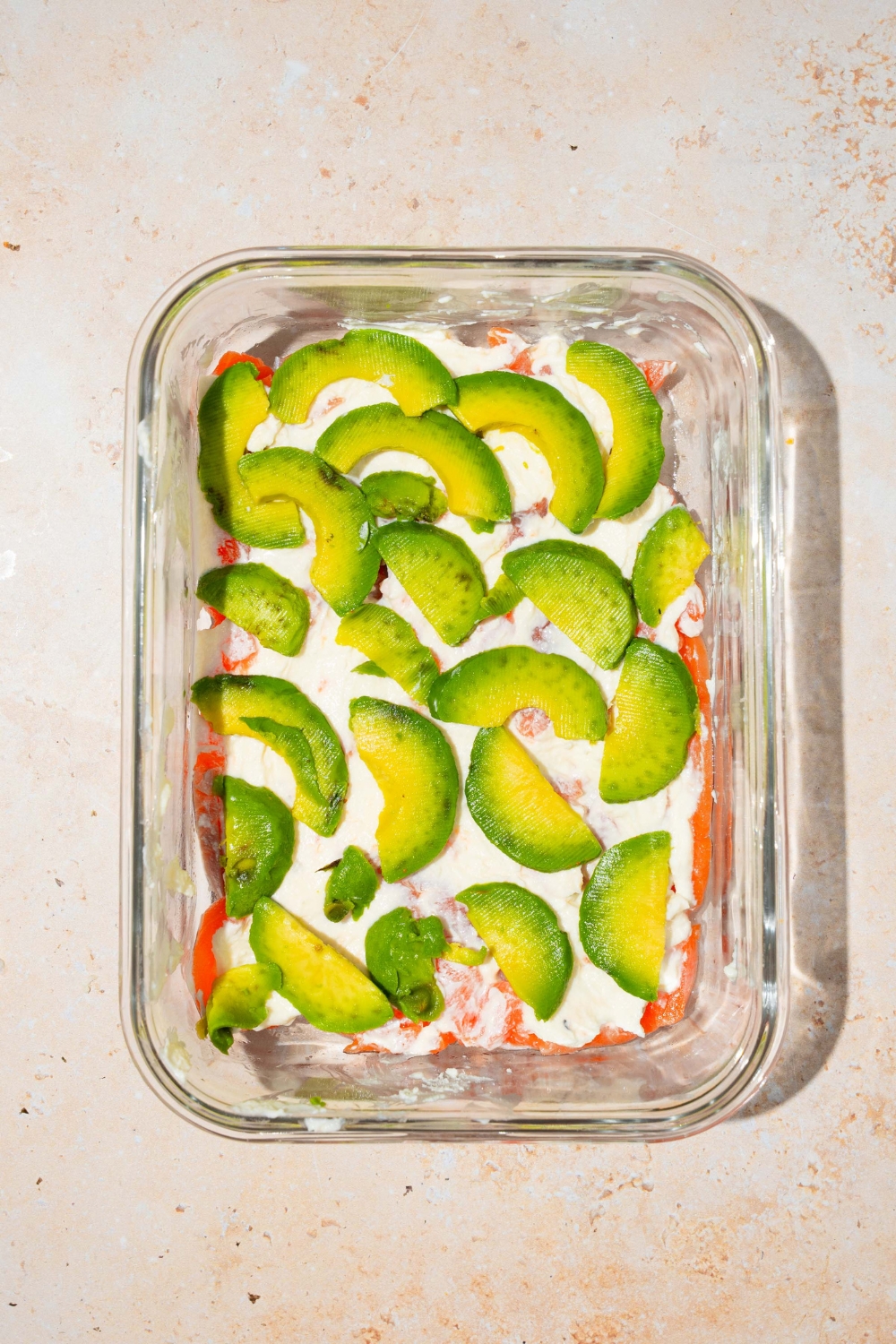 A baking dish with smoked salmon layered with cream cheese and sliced avocado. The dish is on a tan counter.
