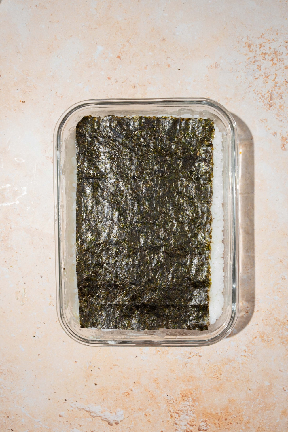 A baking dish with layered lazy sushi including salmon, cream cheese, avocado, rice, and nori. The dish is on a tan counter.
