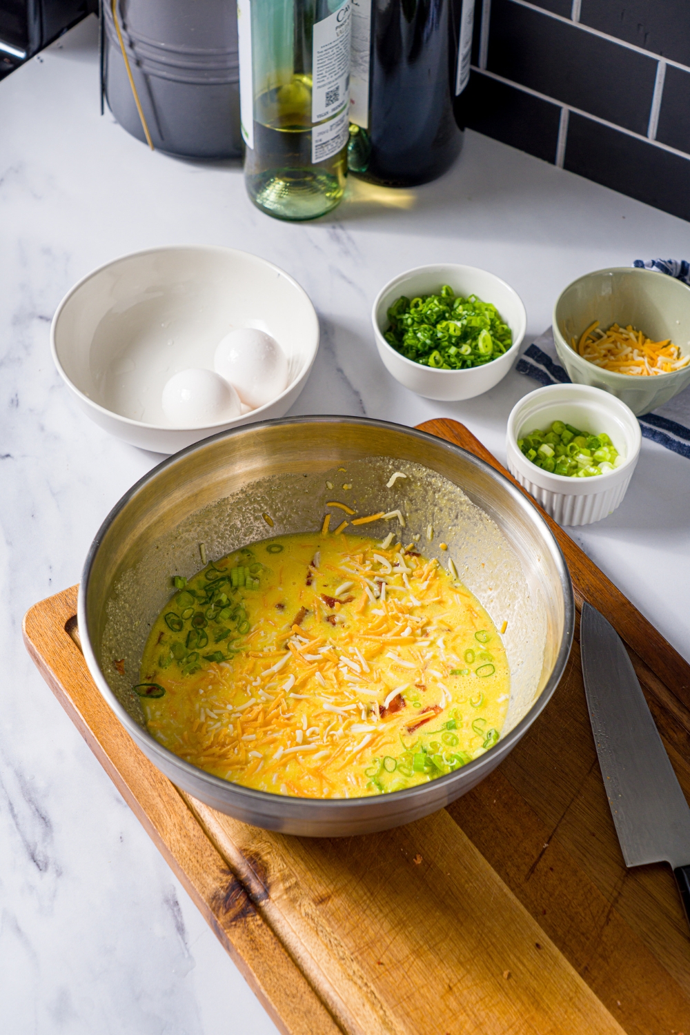 A large bowl with an egg mixture for quiche. The bowl is on a wooden board. The board is on a marble counter with small bowls of ingredients for quiche.