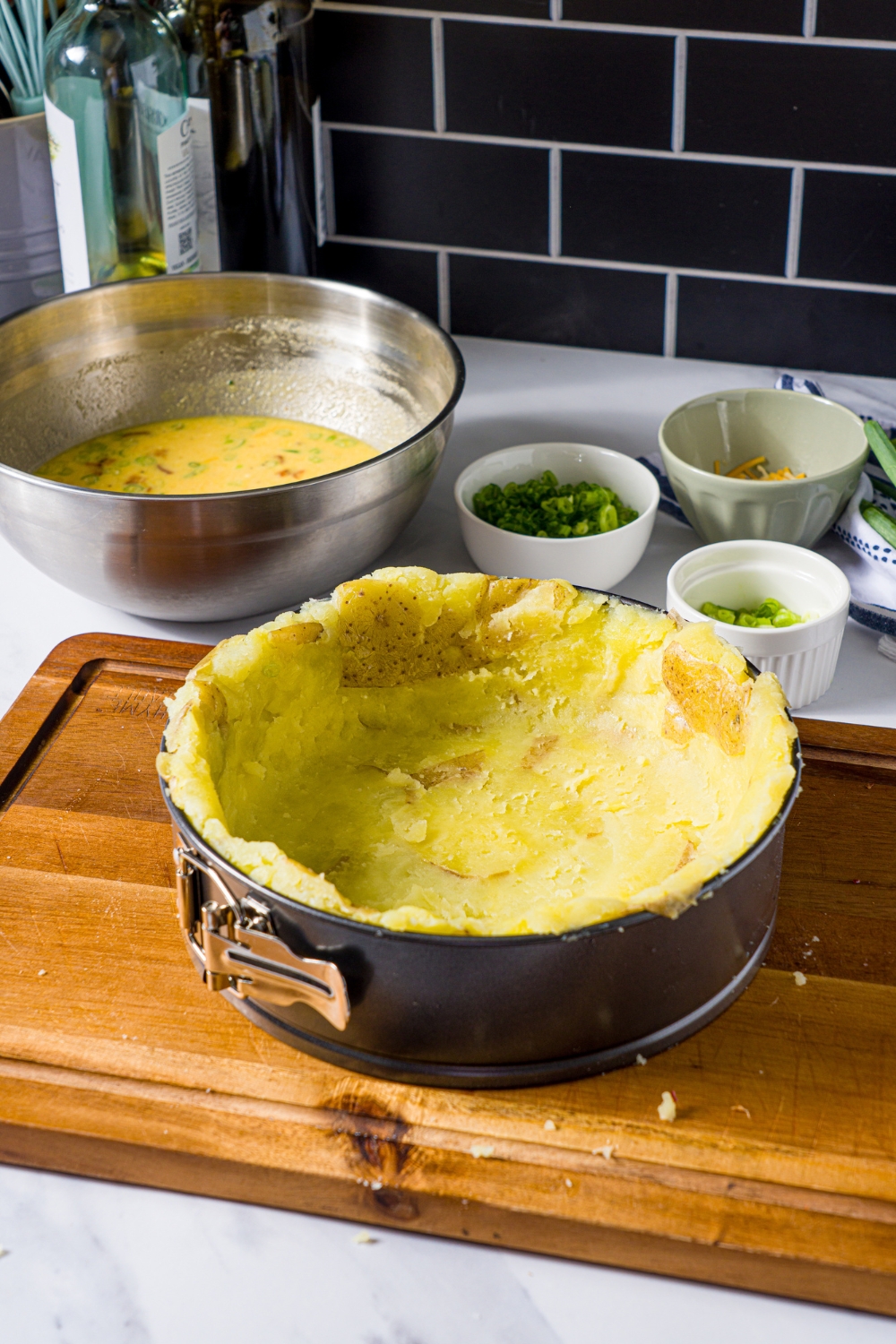A wooden board with a springform pan with shaped potato crust. The board is on a marble counter with bowls of ingredients to make quiche.