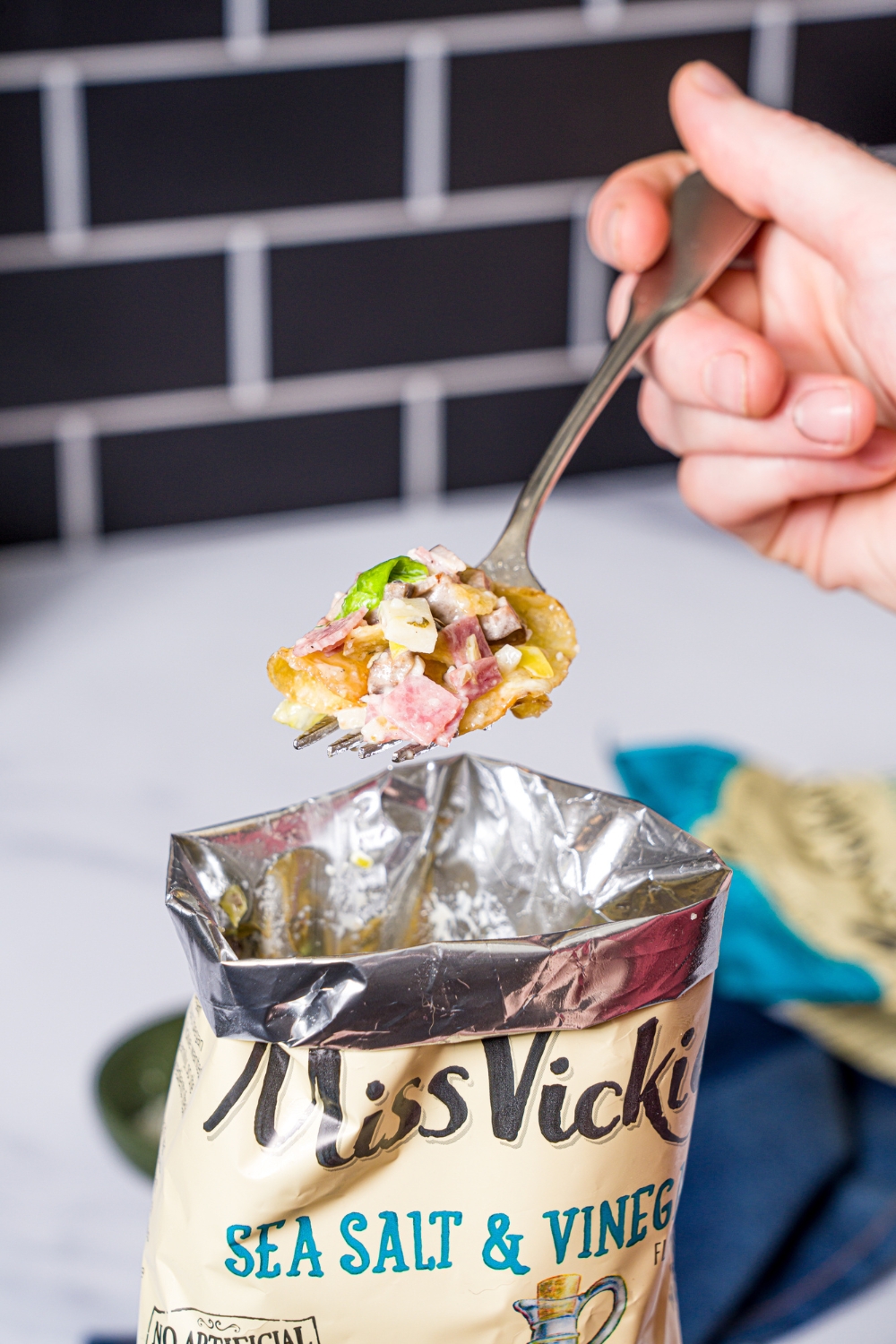 A hand holding a fork that has some salt and vinegar chips and chopped Italian sandwich filling on it, hovering above a bag of salt and vinegar chips.