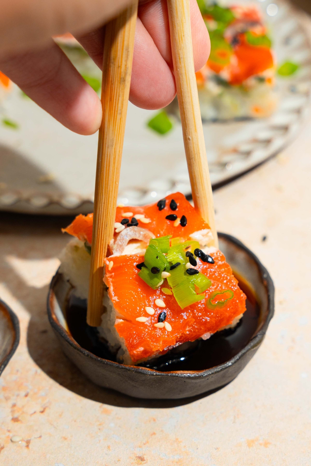 A pair of chopsticks dipping a piece of lazy sushi into a small bowl of soy sauce. The bowl is on a tan counter with a plate of sushi.