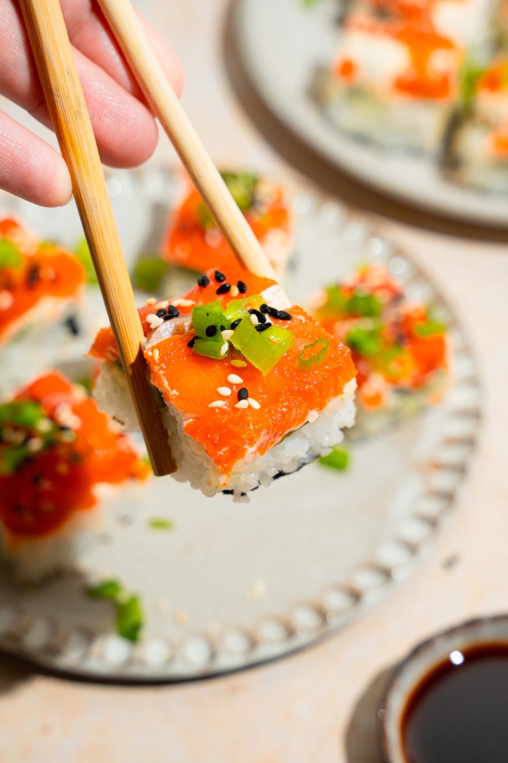 A close up of a pair of chopsticks holding a piece of lazy sushi with salmon topped with sliced green onion and sesame seeds. There is a plate of lazy sushi blurred in the background.