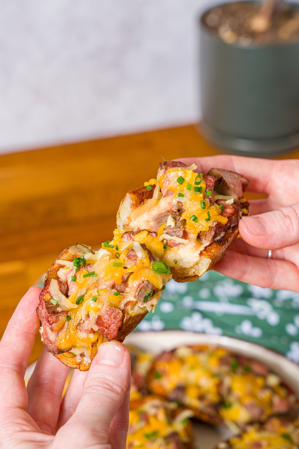 A close up of a hand pulling apart a steak loaded potato skin topped with sliced chives. There is a platter of potato skins and a green floral napkin blurred in the background.