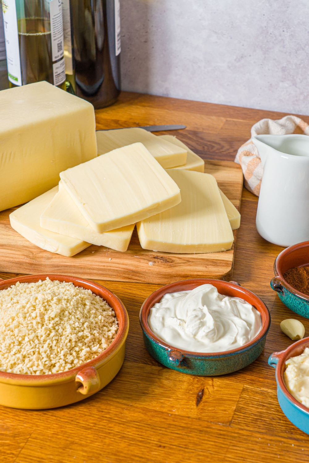 A wooden counter with a ingredietns to make Nashville hot mozzarella sticks including a board of mozzarella cheese sliced into sticks, sour cream, breadcrumbs, and buttermilk.