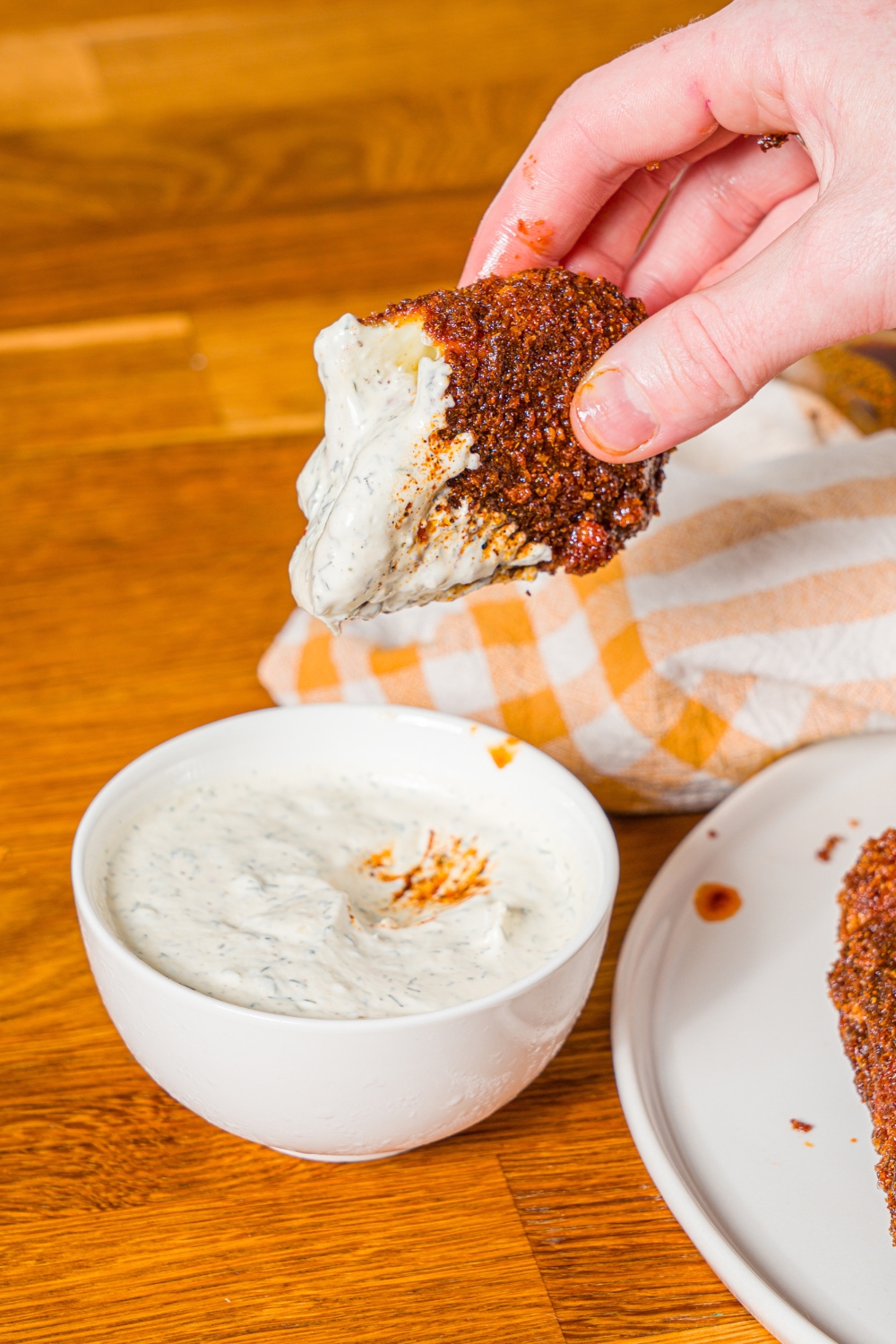 A hand dipping a Nashville hot mozzarella stick into a bowl of ranch. The bowl is on a wooden counter with a plate of mozzarella sticks.