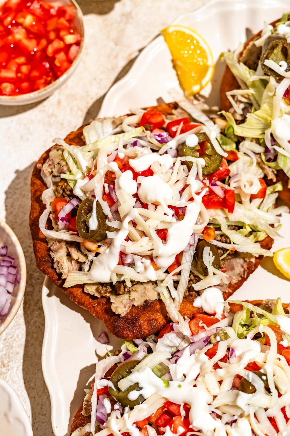 A white platter with several Navajo tacos with Indian fry bread, ground beef, beans, jalapeños, and shredded cabbage with white sauce drizzled on top. The platter is served with sliced lemon.